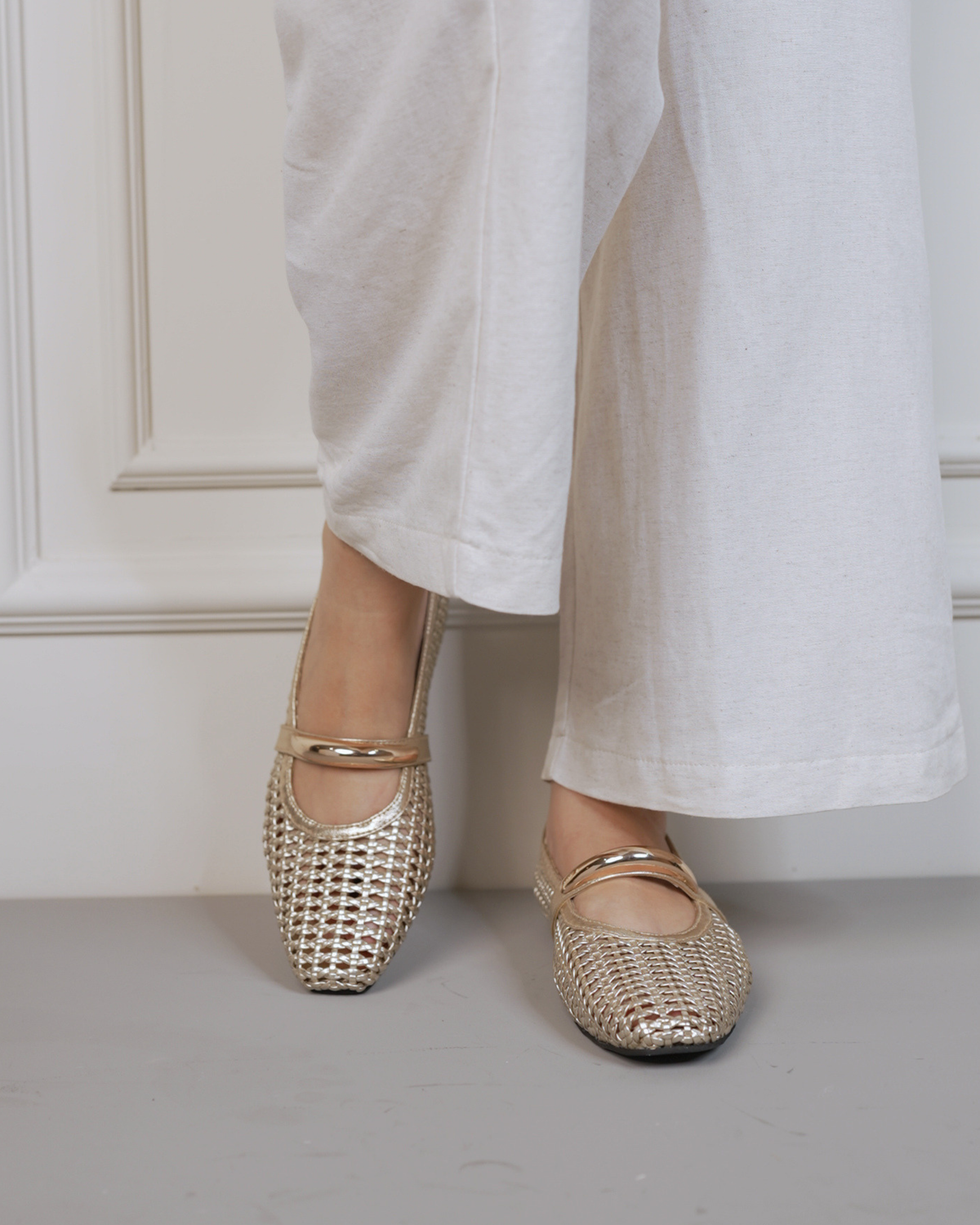 A person wearing white wide-leg trousers and metallic gold woven Mary Jane flats with square toes, standing against a white paneled wall.