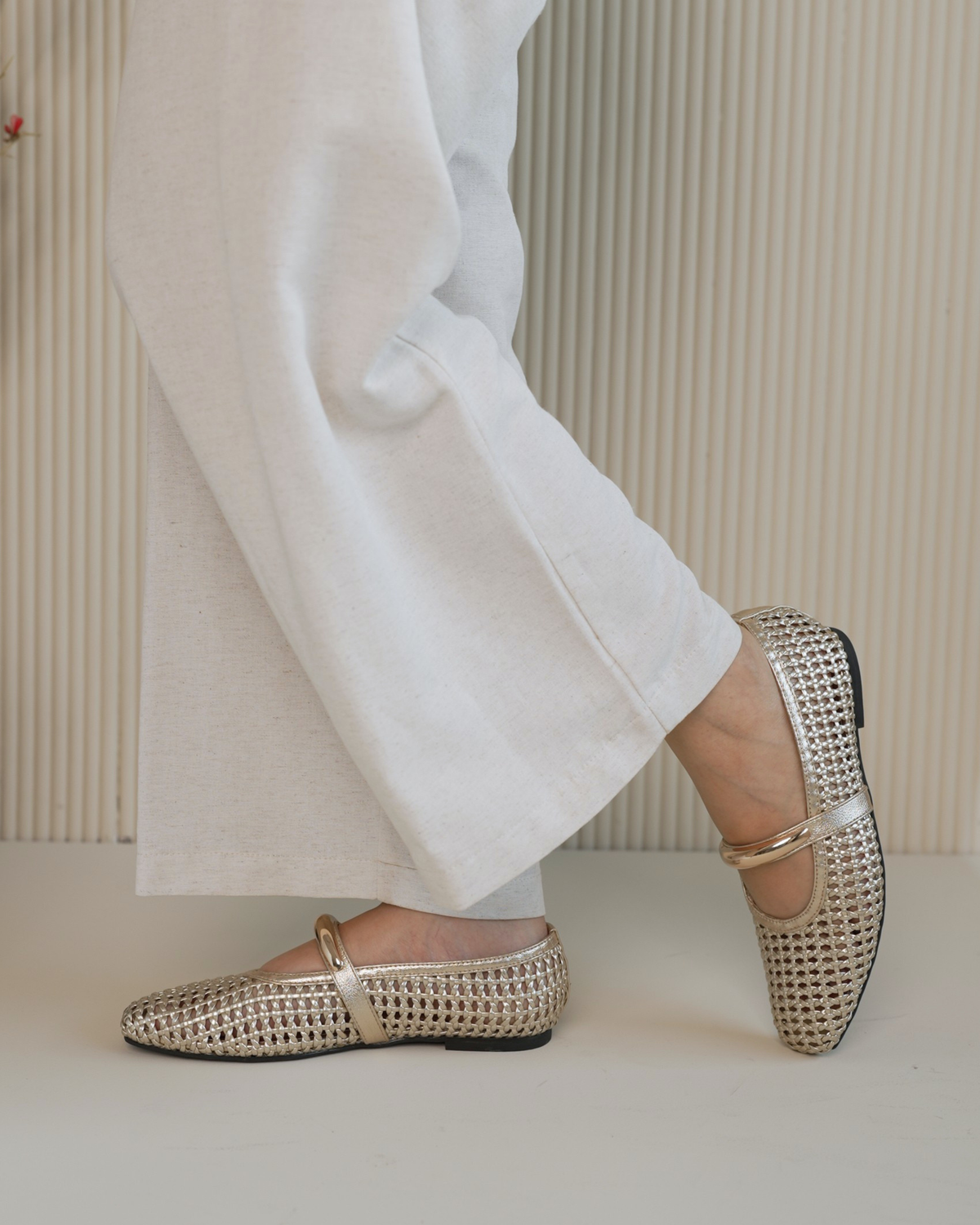 A person wearing white wide-leg pants and metallic gold woven Mary Jane flats, shown from the knees down against a beige ribbed wall and floor.
