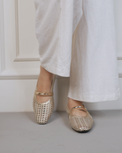 A person wearing white wide-leg trousers and metallic gold woven Mary Jane flats with square toes, standing against a white paneled wall.